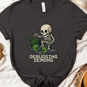 May include: Dark grey t-shirt with a cartoon skeleton wielding a hammer, seated at a laptop displaying green code and two green monster characters. The text "DEBUGGING DEMONS" is below the laptop.