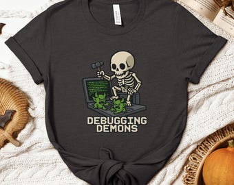 May include: Dark grey t-shirt with a cartoon skeleton wielding a hammer, seated at a laptop displaying green code and two green monster characters. The text "DEBUGGING DEMONS" is below the laptop.