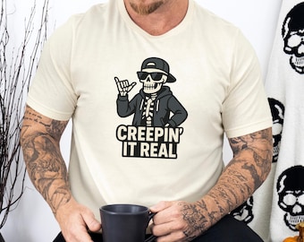 May include: Cream-colored t-shirt featuring a cartoon skeleton wearing sunglasses and a hoodie, making a hand gesture. The text "CREEPIN' IT REAL" is printed below the graphic in a bold, black font. The shirt is a casual, Halloween-themed design.