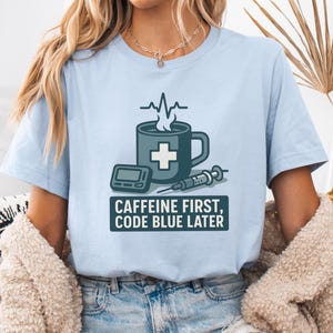 Puede incluir: Camiseta azul claro con un diseño de temática médica. El diseño incluye una taza de café con una cruz, un dispositivo médico y una jeringa. El texto dice "CAFFEINE FIRST, CODE BLUE LATER."