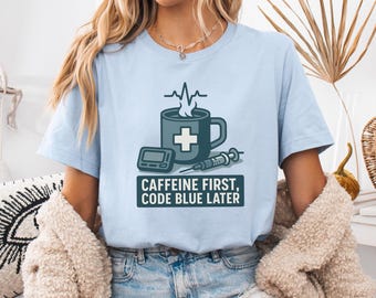 May include: Pale blue t-shirt with a medical-themed design. The graphic includes a coffee mug with a cross, a medical device, and a syringe. The text reads "CAFFEINE FIRST, CODE BLUE LATER."