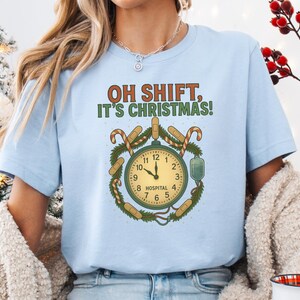 Könnte beinhalten: Hellblaues T-Shirt mit dem Text "OH SHIFT, IT'S CHRISTMAS!". Die Grafik zeigt eine Uhr, umgeben von Zuckerstangen, Pflastern und einem Infusionsbeutel. Die Uhrzeiger zeigen auf 12:00 Uhr.