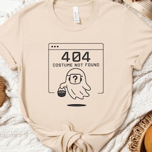 May include: A beige t-shirt with a black graphic. The graphic shows a ghost with a question mark inside a browser window, and the text "404 COSTUME NOT FOUND". The shirt has rolled sleeves and a round neckline.