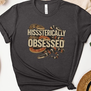 Op de afbeelding: Donkergrijs t-shirt met de tekst "Hissssterically Obsessed" en illustraties van slangen in bruin, oranje en zwart. Het shirt heeft een ronde hals en korte mouwen.