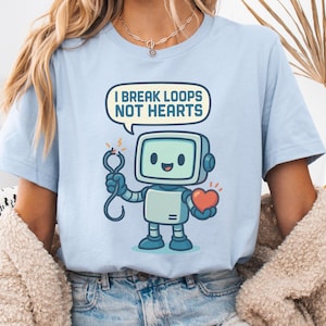 May include: Pale blue t-shirt with a cartoon robot design. The robot, with a screen face, holds a heart and a tool, and a speech bubble says "I BREAK LOOPS NOT HEARTS". The shirt is made of a soft material.
