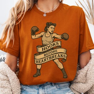 May include: Rust-colored t-shirt featuring a vintage illustration of a female boxer in a fighting stance, wearing boxing gloves. A banner across the image reads "HOOKS BEFORE HEARTBREAKS". The design is a retro-style graphic.