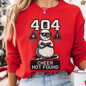 May include: A red sweatshirt with a snowman graphic wearing sunglasses and crossed arms. The text "404 CHEER NOT FOUND" is displayed above and below the snowman. Snowflake and warning symbols are also present. The sweatshirt is a festive holiday design.