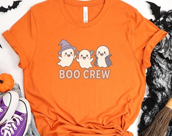 Può includere: T-shirt arancione con tre fantasmi dei cartoni animati e la scritta "BOO CREW". I fantasmi indossano un cappello da strega, un'ala di pipistrello e un mantello. Perfetta per Halloween.