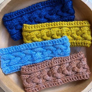 May include: Four rectangular headbands featuring a chunky cable knit pattern. The headbands are colored in deep navy blue, mustard yellow, light sky blue, and a muted terracotta brown, each showcasing a thick, textured braided design.