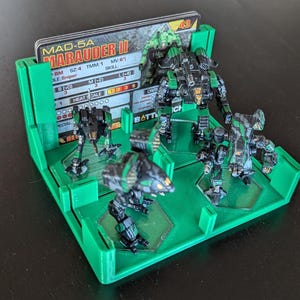 May include: A green plastic game organiser with four compartments for miniature figures. Each compartment has a clear plastic insert and a card slot. The card shows the text "MAD-5A MARAUDER II" and has stats for a game piece.