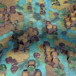 May include: A tabletop game board featuring a hexagonal grid layout with raised brown terrain tiles and flat blue water sections. The board includes elevated brown hexagonal pillars with green textured tops, creating a multi-level landscape.