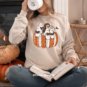 May include: Beige sweatshirt featuring a Halloween-themed graphic. The design includes a pumpkin made of books with ghosts reading. The sweatshirt has a crew neck and long sleeves. The text "@MemoryArt666" is visible.