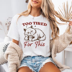 May include: White t-shirt with a graphic of a grumpy cat holding a coffee cup, and the text "TOO TIRED For This" in maroon. The cat is drawn in a relaxed pose, with a bored expression. The shirt is a casual wear item.