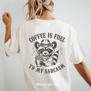 Puede incluir: Una camiseta blanca con una ilustraci&oacute;n en blanco y negro de un mapache con un sombrero de vaquero y sosteniendo una taza de caf&eacute;. El texto "COFFEE IS FUEL TO MY SARCASM" est&aacute; impreso en la camiseta. El texto "@MemoryArt666" est&aacute; impreso en la parte inferior de la camiseta.