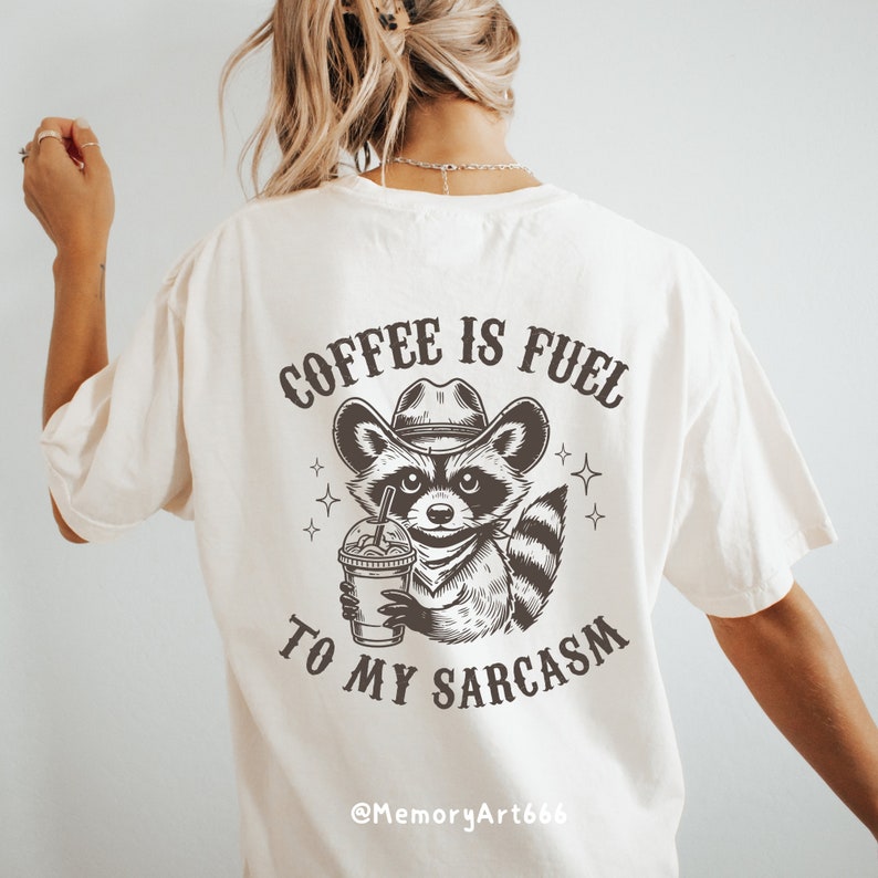 Puede incluir: Una camiseta blanca con una ilustraci&oacute;n en blanco y negro de un mapache con un sombrero de vaquero y sosteniendo una taza de caf&eacute;. El texto "COFFEE IS FUEL TO MY SARCASM" est&aacute; impreso en la camiseta. El texto "@MemoryArt666" est&aacute; impreso en la parte inferior de la camiseta.