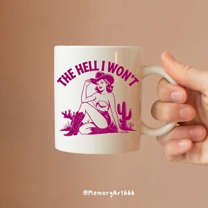 May include: White ceramic mug with a pink graphic of a woman in a cowboy hat sitting with a cactus. The text "THE HELL I WON'T" is written around the woman.