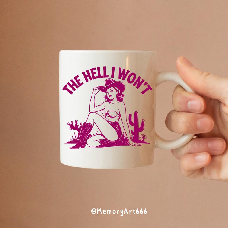 May include: White ceramic mug with a pink graphic of a woman in a cowboy hat sitting with a cactus. The text "THE HELL I WON'T" is written around the woman.