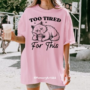 May include: A light pink t-shirt with the text "TOO TIRED For This" in black, featuring a grumpy cat graphic. The cat is drawn in black and white, resting its head on its paw and holding a coffee cup. The shirt is a casual, oversized fit.