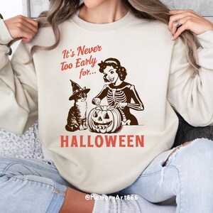Puede incluir: Una sudadera blanca con el texto "It's Never Too Early for... HALLOWEEN" en rojo y marr&oacute;n. Una mujer de dibujos animados con un cuerpo de esqueleto est&aacute; tallando una calabaza con un gato que lleva un sombrero de bruja a su lado.