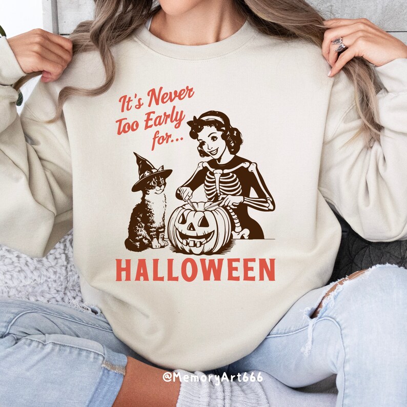 Puede incluir: Una sudadera blanca con el texto "It's Never Too Early for... HALLOWEEN" en rojo y marr&oacute;n. Una mujer de dibujos animados con un cuerpo de esqueleto est&aacute; tallando una calabaza con un gato que lleva un sombrero de bruja a su lado.