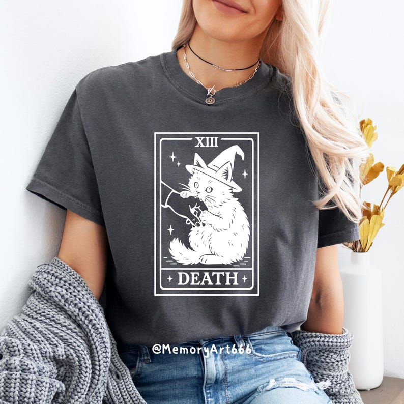 May include: Dark gray t-shirt featuring a white graphic of a cat wearing a witch hat, labeled "DEATH" with the Roman numeral "XIII". The cat holds a scythe. The shirt is a comfortable, casual style.