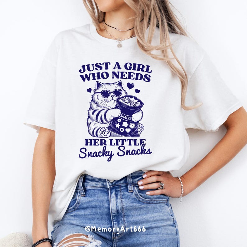 May include: White t-shirt with a blue graphic of a cat wearing sunglasses and holding a bag of snacks. The text on the shirt reads "Just a girl who needs her little snacky snacks".