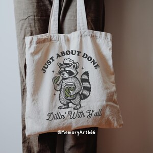 K&ouml;nnte beinhalten: Cremefarbene Tragetasche mit einem Waschb&auml;ren, der einen Cowboyhut tr&auml;gt und ein Glas Gurken h&auml;lt. Der Text auf der Tasche lautet "JUST ABOUT DONE" und "Dillin' With Y'all". Die Tasche hat zwei Griffe und ist aus weichem Stoff.