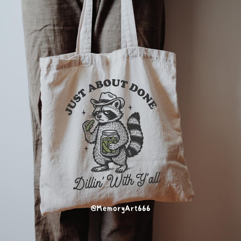 K&ouml;nnte beinhalten: Cremefarbene Tragetasche mit einem Waschb&auml;ren, der einen Cowboyhut tr&auml;gt und ein Glas Gurken h&auml;lt. Der Text auf der Tasche lautet "JUST ABOUT DONE" und "Dillin' With Y'all". Die Tasche hat zwei Griffe und ist aus weichem Stoff.