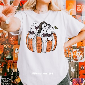 May include: White t-shirt featuring a Halloween-themed graphic. The design shows four ghosts reading books inside a pumpkin made of books. The pumpkin is orange and the books are white and orange. The text "@MemoryArt666" is at the bottom.