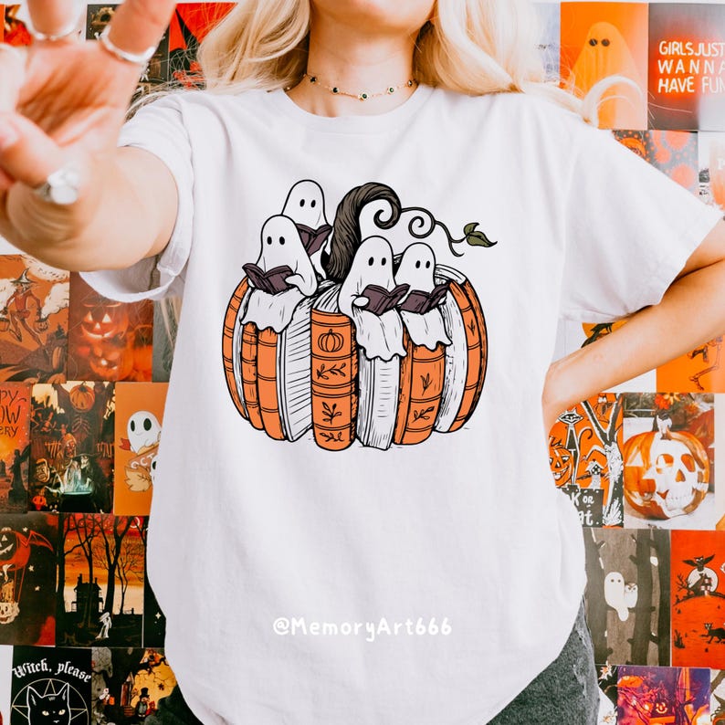 May include: White t-shirt featuring a Halloween-themed graphic. The design shows four ghosts reading books inside a pumpkin made of books. The pumpkin is orange and the books are white and orange. The text "@MemoryArt666" is at the bottom.