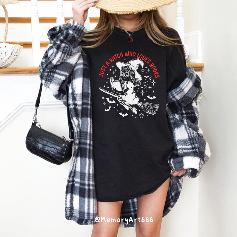 Może przedstawiać: Czarny T-shirt z białym nadrukiem przedstawiającym czarownicę latającą na miotle i czytającą książkę. Tekst "Just a witch who loves books" jest napisany na czerwono wok&oacute;ł grafiki.