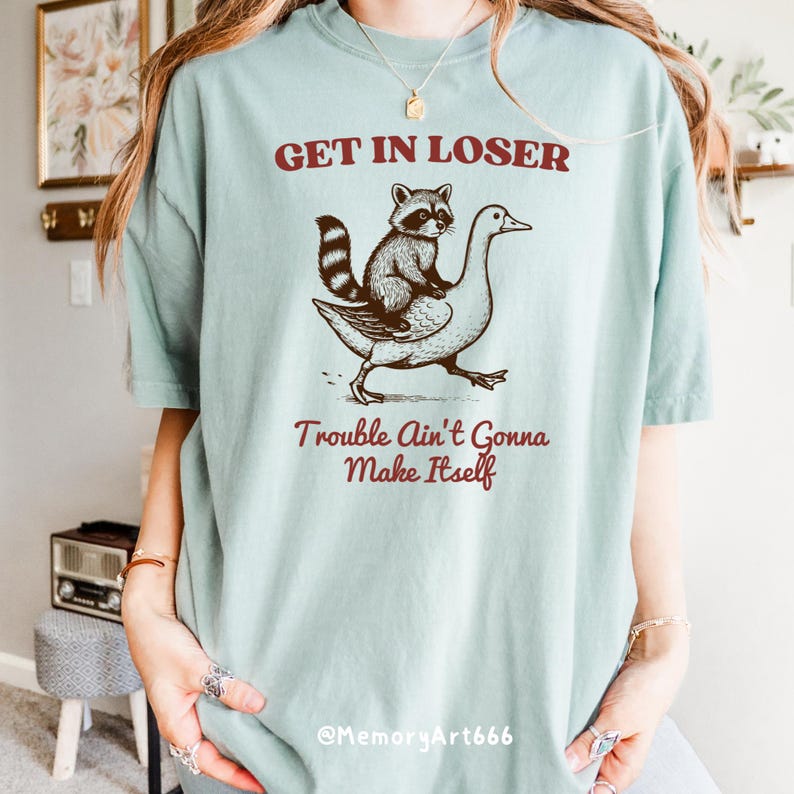 May include: A light blue t-shirt with a graphic of a raccoon riding a goose. The text "Get in Loser, Trouble Ain't Gonna Make Itself" is printed in red on the shirt.