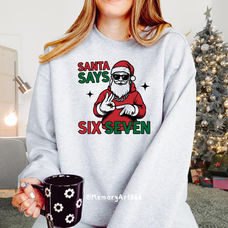May include: A light gray sweatshirt with a graphic of Santa Claus wearing sunglasses and a red suit. The text "SANTA SAYS SIX SEVEN" is printed above and below the Santa graphic in red and green. A black mug with white floral designs is held in the person's hand.
