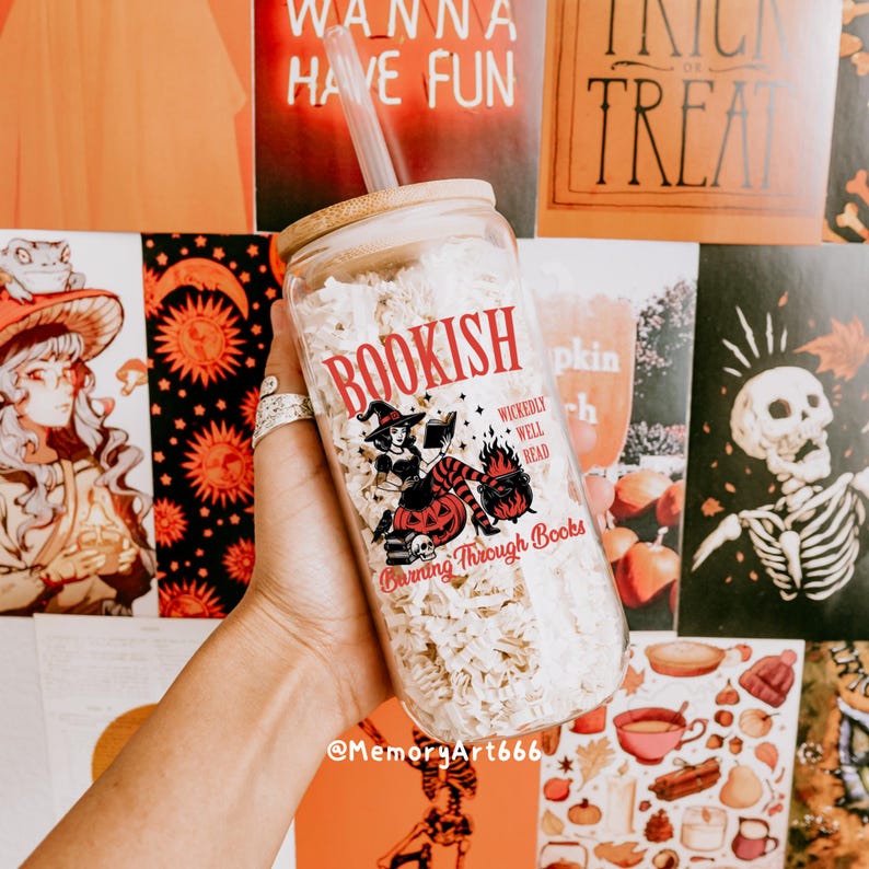 May include: A clear glass jar with a bamboo lid and straw, filled with white material. The jar features the word "BOOKISH" in red, along with a witch illustration and the text "Burning Through Books". The background includes Halloween-themed posters.