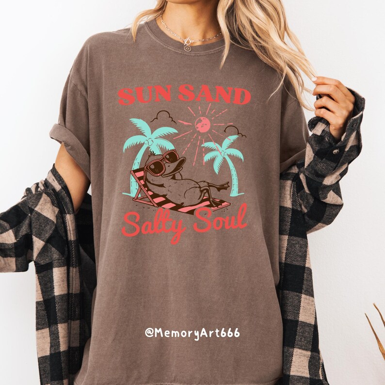 Peut inclure: Un t-shirt marron avec un motif graphique repr&eacute;sentant un canard de dessin anim&eacute; portant des lunettes de soleil et se relaxant sur une chaise de plage sous des palmiers. Le texte "Sun Sand Salty Soul" est imprim&eacute; sur le t-shirt. Le texte "@MemoryArt666" est imprim&eacute; en bas du t-shirt.