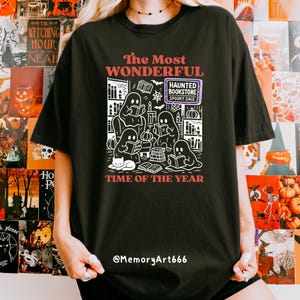 K&ouml;nnte beinhalten: Schwarzes T-Shirt mit einer wei&szlig;en Grafik, die Geister beim Lesen von B&uuml;chern, eine Katze und den Text "The Most Wonderful Time of the Year" zeigt. Das Design enth&auml;lt den Text "Haunted Bookstore Spooky Sale".
