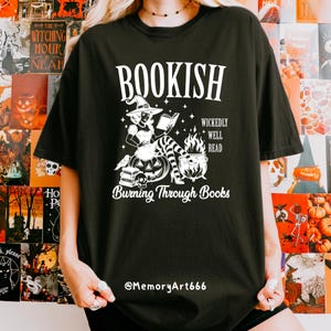 May include: Black t-shirt with a white graphic featuring a witch reading a book, a cauldron, and the text "BOOKISH". Additional text includes "WICKEDLY WELL READ" and "Burning Through Books". The shirt also has the text "@MemoryArt666".