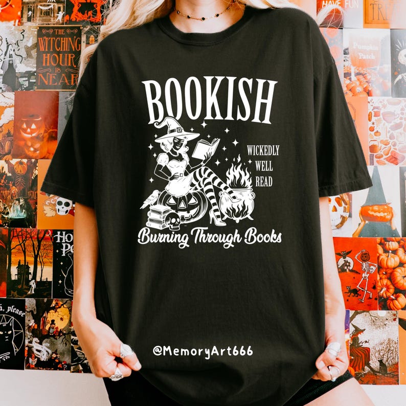 May include: Black t-shirt with a white graphic featuring a witch reading a book, a cauldron, and the text "BOOKISH". Additional text includes "WICKEDLY WELL READ" and "Burning Through Books". The shirt also has the text "@MemoryArt666".