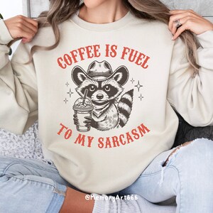 Puede incluir: Una sudadera beige con el texto "Coffee is fuel to my sarcasm" en rojo. Un mapache de dibujos animados con un sombrero de vaquero y sosteniendo una taza de caf&eacute; est&aacute; en el centro del texto.