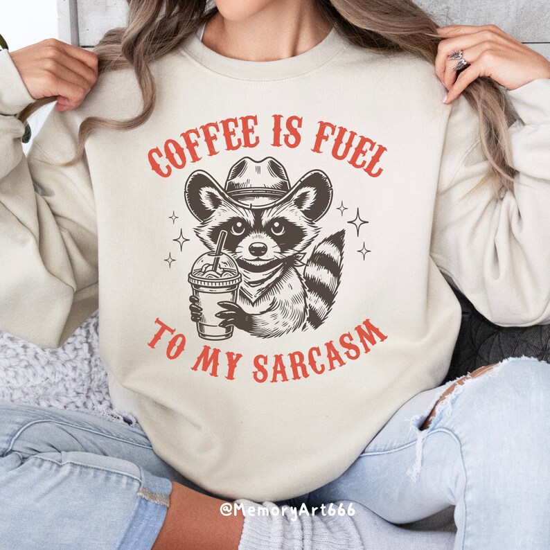 Puede incluir: Una sudadera beige con el texto "Coffee is fuel to my sarcasm" en rojo. Un mapache de dibujos animados con un sombrero de vaquero y sosteniendo una taza de caf&eacute; est&aacute; en el centro del texto.