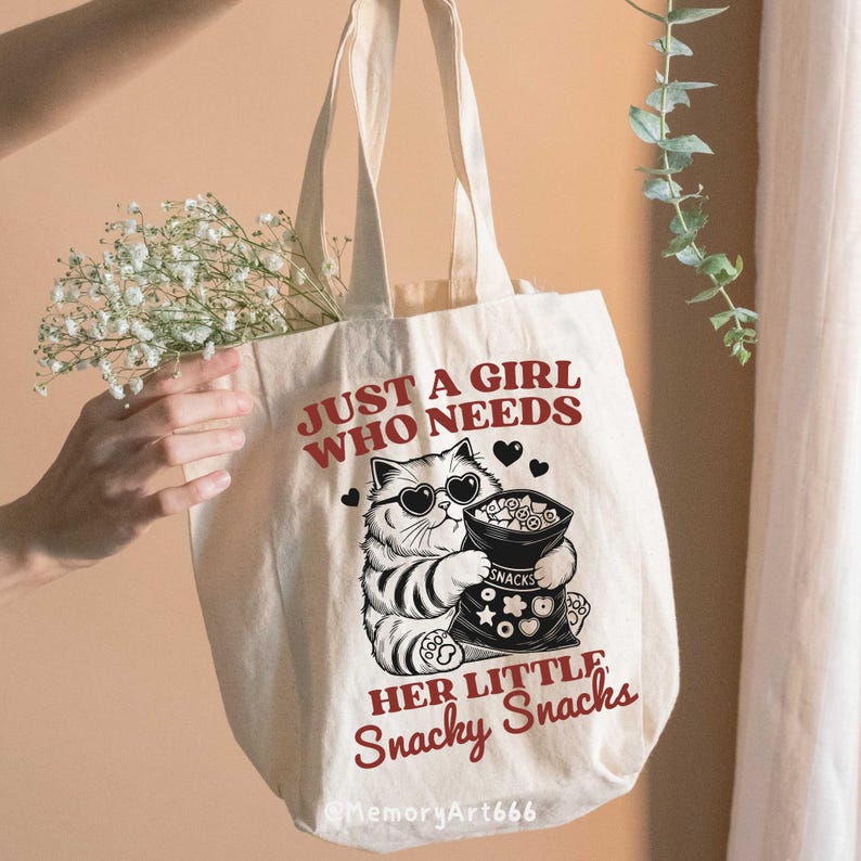 May include: A white canvas tote bag with the text "Just a girl who needs her little snacky snacks" in red. A cartoon cat wearing sunglasses holds a bag of snacks.