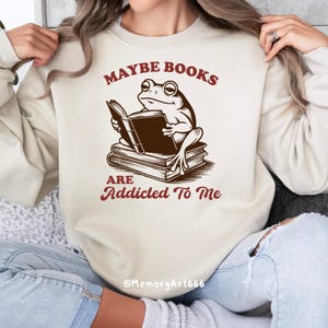 May include: Beige sweatshirt with a frog reading a book. The frog is sitting on a stack of books. The text "MAYBE BOOKS ARE ADDICTED TO ME" is printed in maroon.