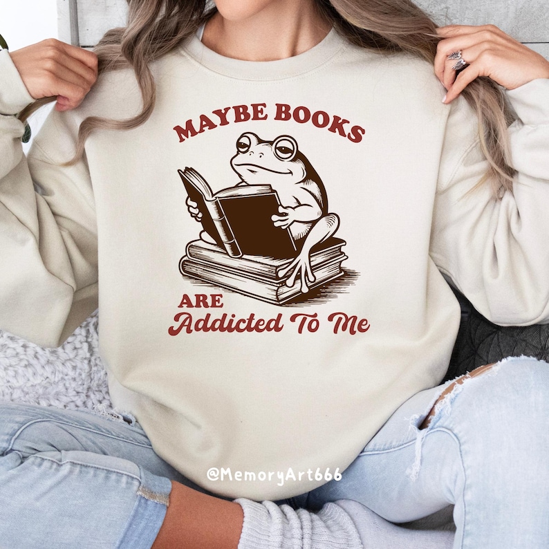 May include: Beige sweatshirt with a frog reading a book. The frog is sitting on a stack of books. The text "MAYBE BOOKS ARE ADDICTED TO ME" is printed in maroon.