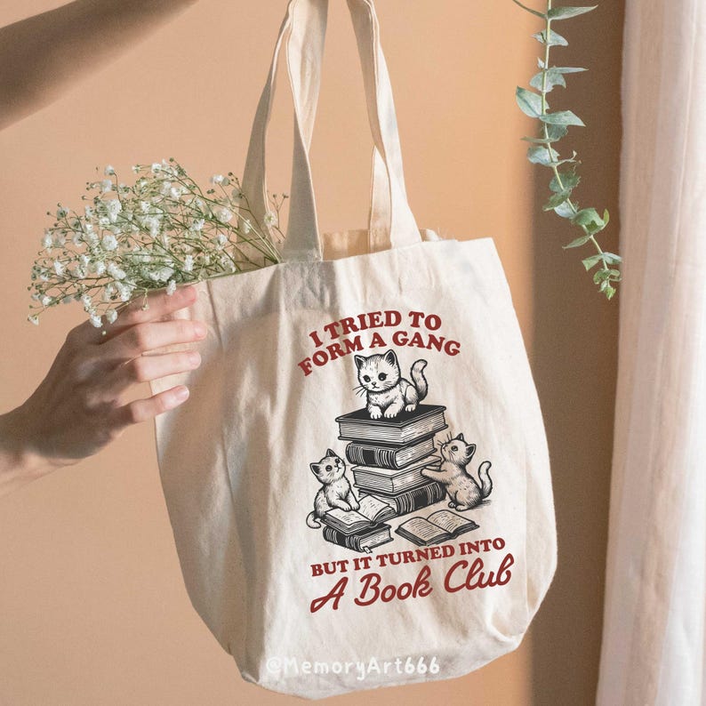 May include: A white tote bag with a red and black graphic of cats reading books. The text on the bag reads "I tried to form a gang but it turned into a book club".