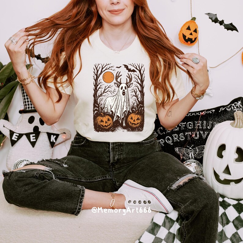 May include: Cream-colored t-shirt featuring a Halloween-themed graphic. The design includes a ghost, pumpkins, trees, and a deer with antlers, set against a full moon. The shirt is paired with black jeans and white sneakers.