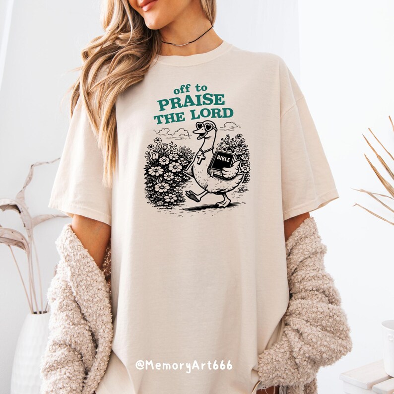 Op de afbeelding: Cr&egrave;mekleurig T-shirt met de tekst "off to PRAISE THE LORD" in teal. De afbeelding toont een cartoon gans met een zonnebril en een kruis ketting, die een Bijbel draagt. De gans loopt door een bloemenveld.