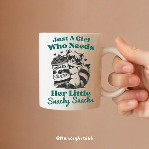 May include: White ceramic mug with a green and black design. The mug features a raccoon holding a bag of snacks with the text "Just a girl who needs her little snacky snacks".