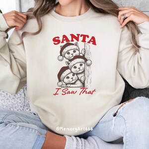 May include: A cream coloured sweatshirt with a red graphic of three cats wearing Santa hats peeking around a corner. The text "SANTA I Saw That" is printed below the cats. The text "@MemoryArt666" is printed at the bottom of the sweatshirt.
