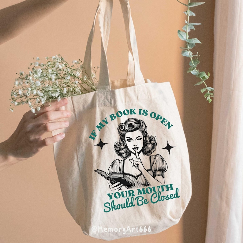 May include: A white tote bag with a black and white illustration of a woman with her finger to her lips. The text on the bag reads "If my book is open your mouth should be closed".