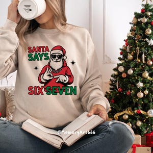 May include: Beige sweatshirt with a graphic of Santa Claus wearing sunglasses and a red outfit. The text "SANTA SAYS SIX SEVEN" is printed above and below the Santa graphic. A Christmas tree is in the background.